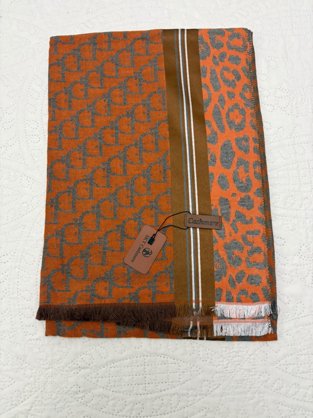 An orange woven scarf with a gray repeating monogram pattern, a leopard print section, and a vertical brown stripe with white lines, laid flat on a white textured background with a brand tag attached.