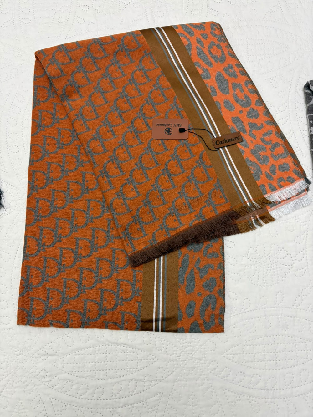 An orange and gray patterned scarf with a brown and gray leopard print section, featuring a monogram pattern and a fringed edge, displayed on a white textured background.