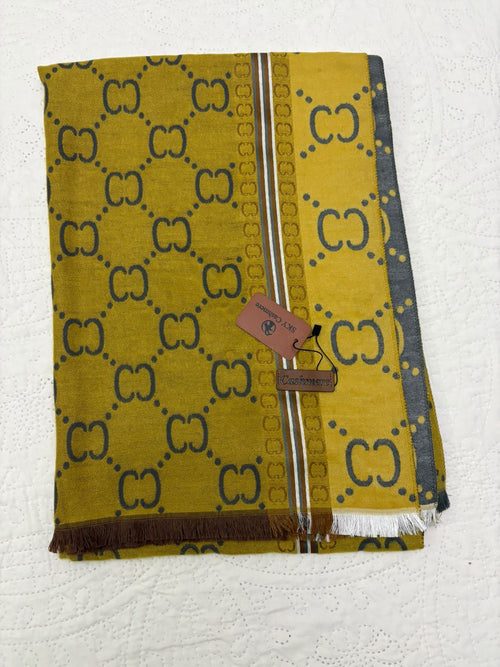 A folded Gucci scarf in a mustard yellow color with a dark brown GG monogram pattern, featuring a brown leather brand tag and fringed edges, laid flat on a white textured background.
