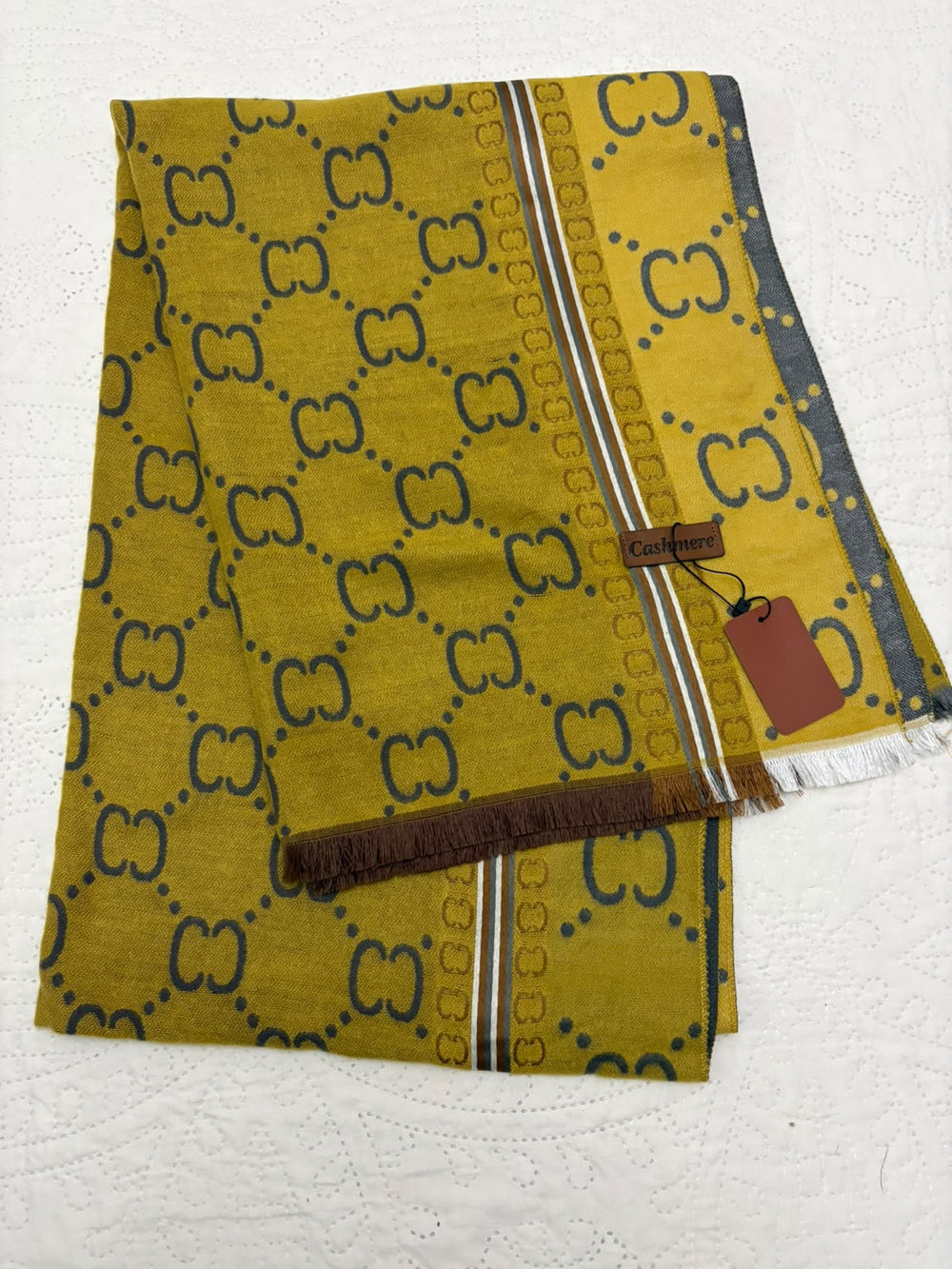 A folded yellow scarf featuring a dark blue repeating interlocking monogram pattern, with a striped border and fringed edges, shown on a white textured background with a brown brand tag attached.
