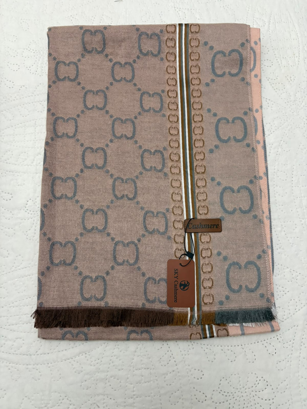 A folded Gucci scarf in a dusty pink color with a blue monogram pattern and a central gold chain motif, featuring a brown leather brand tag.