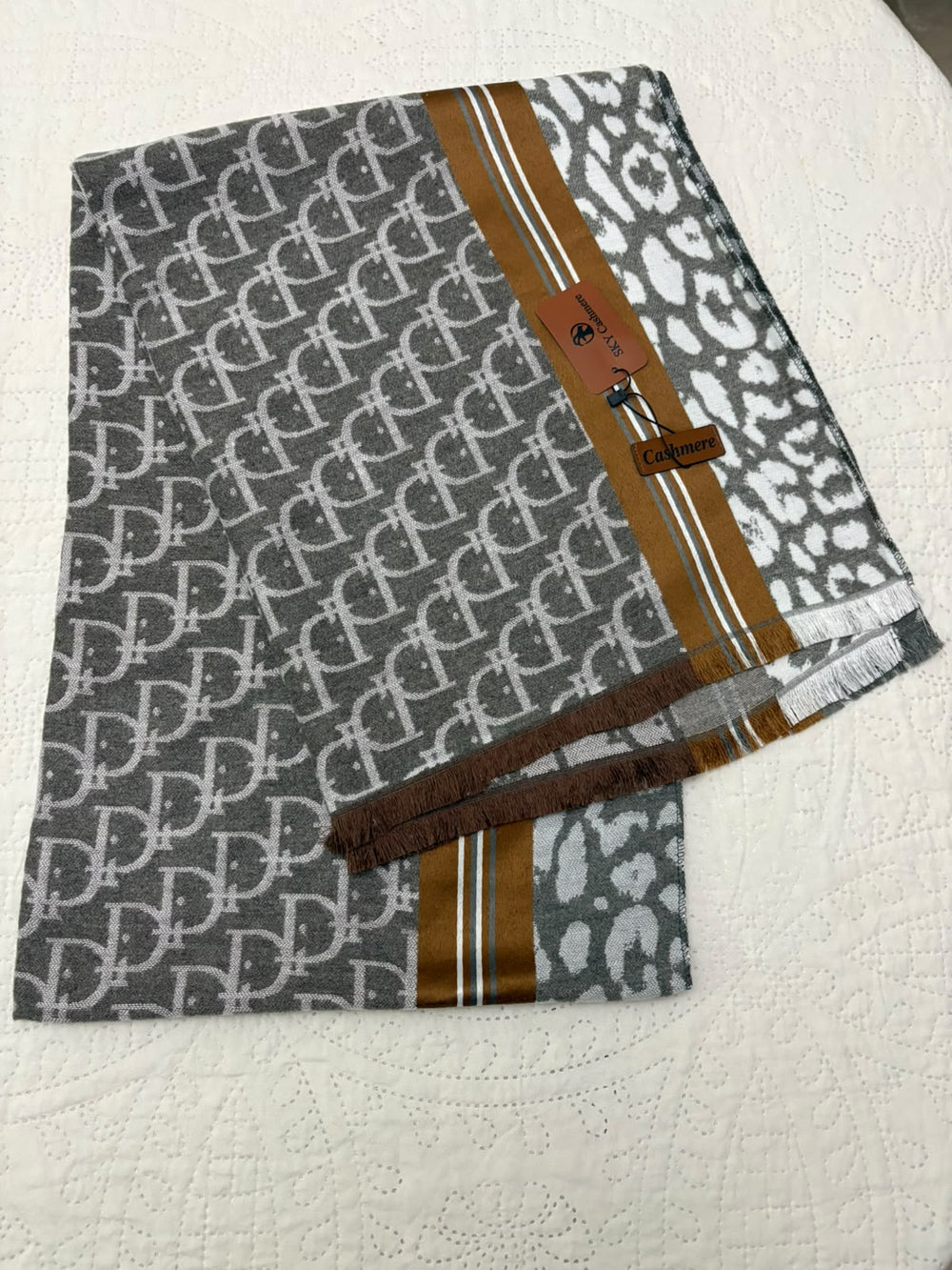 A folded designer scarf featuring a gray and white interlocking monogram pattern, a brown and white leopard print section, and a central brown stripe with gold lines. A small brown leather tag with a logo is attached near the top right.