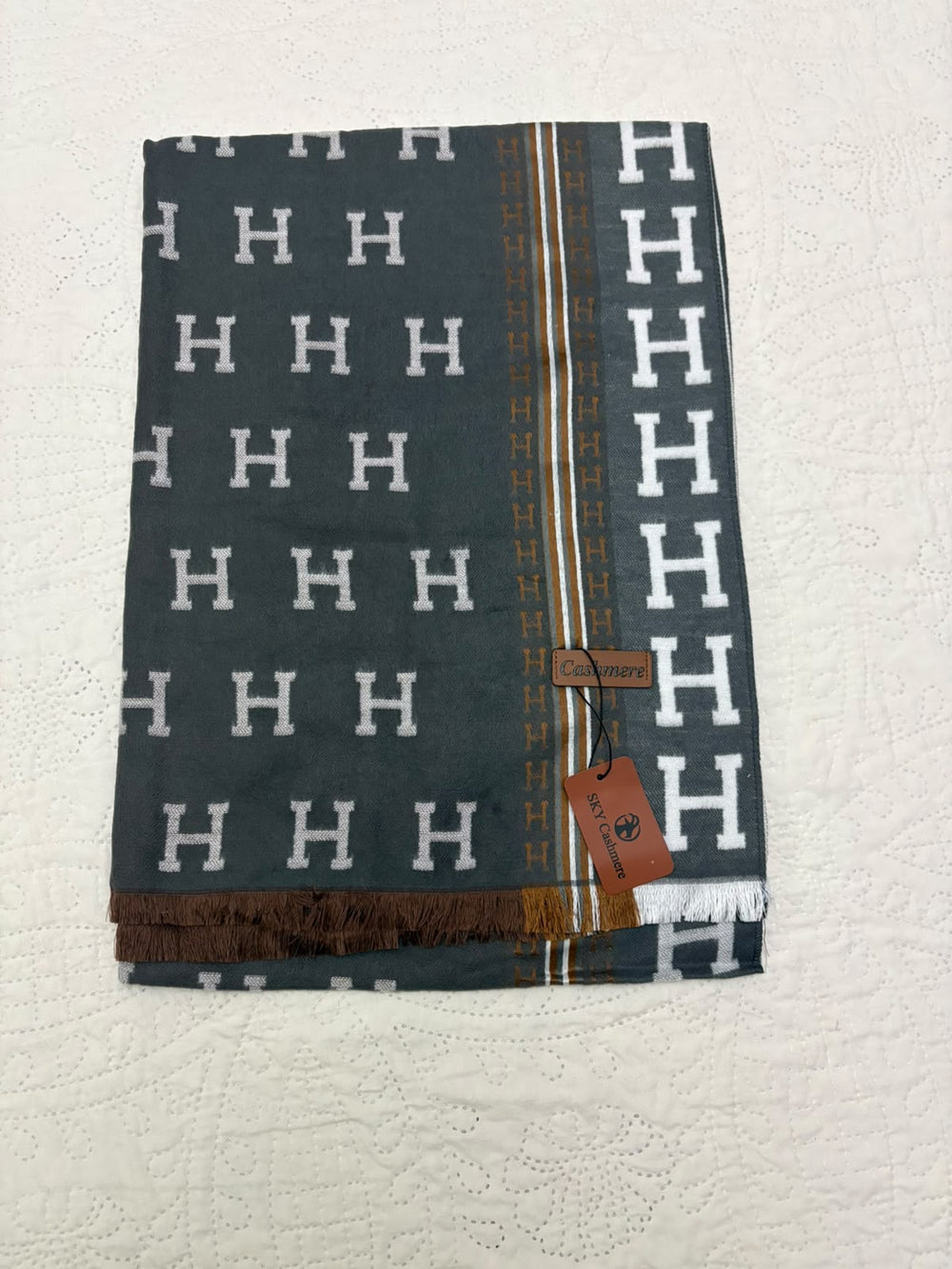 A folded Hermès scarf in dark gray with a white H logo pattern, featuring a vertical gold stripe with brown accents and a brown leather brand tag.