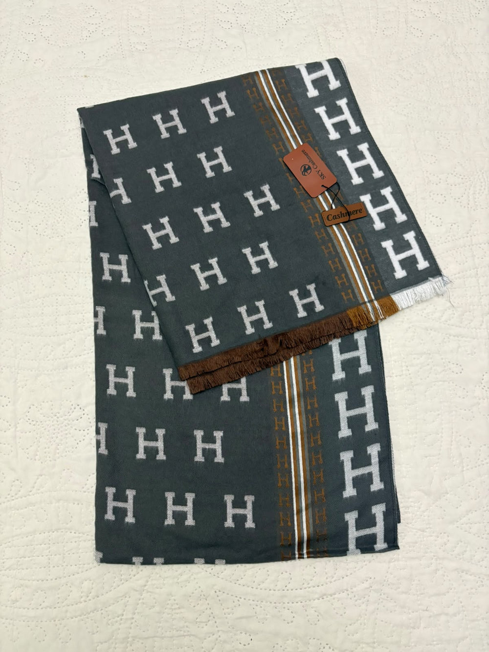 A folded dark gray scarf featuring a repeating white 'H' pattern, a brown and gold striped border, and a small brown leather brand tag, displayed on a textured off-white background.