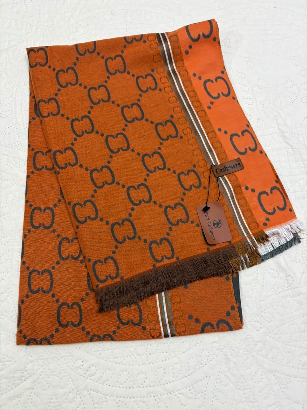 An orange Gucci scarf with a dark brown GG monogram pattern, featuring a striped border and fringed edges, laid flat on a white textured background.
