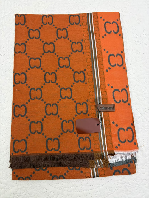 An orange Gucci scarf featuring the signature GG monogram pattern in dark brown, with a striped border and fringed edges, laid flat on a white textured background.