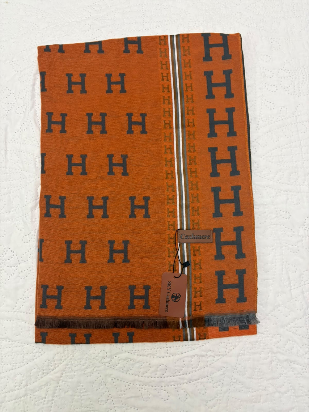 An orange woven scarf with a repeating navy blue 'H' pattern, featuring a central vertical stripe with 'H' motifs and a small brown leather Hermès tag, laid flat on a white textured fabric background.