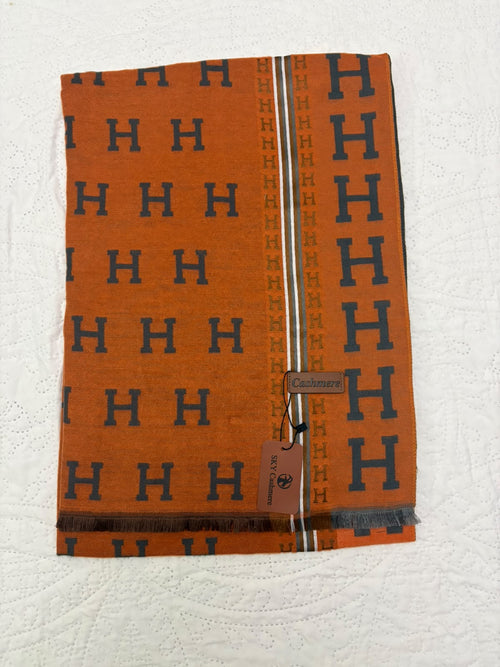 An orange woven scarf with a repeating navy blue 'H' pattern, featuring a central vertical stripe with 'H' motifs and a small brown leather Hermès tag, laid flat on a white textured fabric background.