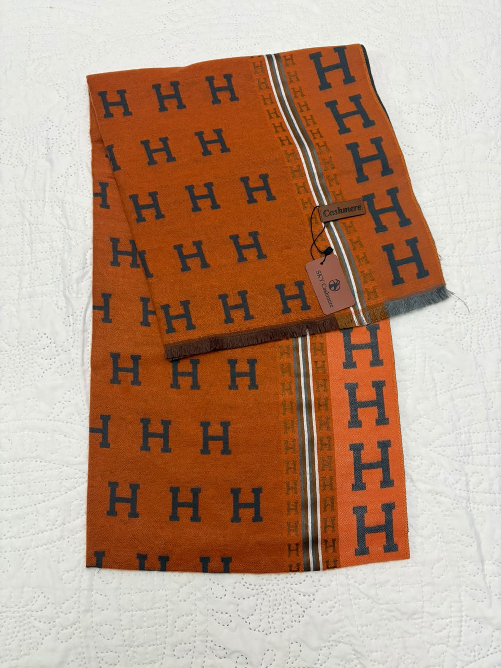 An orange woven scarf with a repeating navy blue 'H' monogram pattern and a striped border, laid flat on a white textured fabric background. A small brand tag is attached.