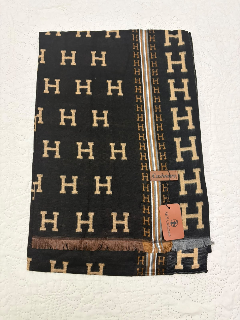 A folded black knit scarf with a repeating beige 'H' pattern, featuring a central vertical stripe with 'HH' motifs and a fringed hem. A small orange tag is attached, and the scarf is laid flat on a textured off-white surface.