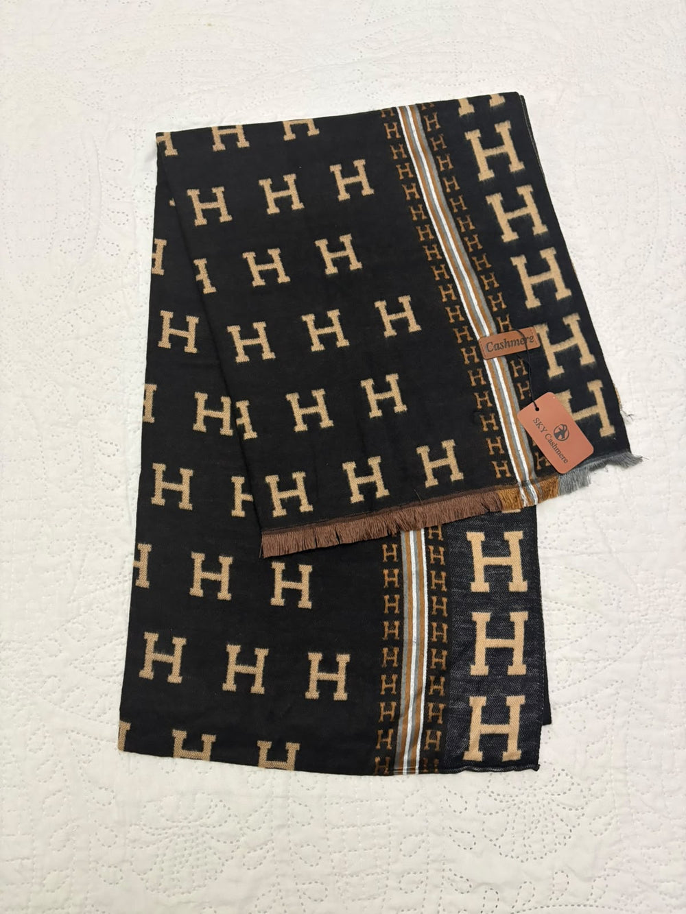 A folded black scarf featuring a repeating beige 'H' pattern and a striped border with beige, white, and brown details, displayed on a textured white surface.