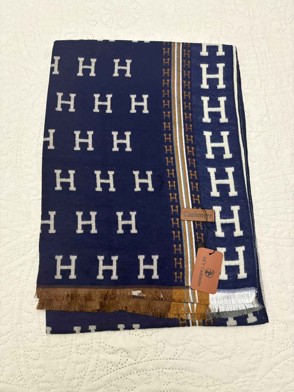 A folded navy blue scarf with a repeating white 'H' pattern, gold stripes, and fringed edges, displayed on a textured off-white surface. A brown leather tag is attached to the scarf.