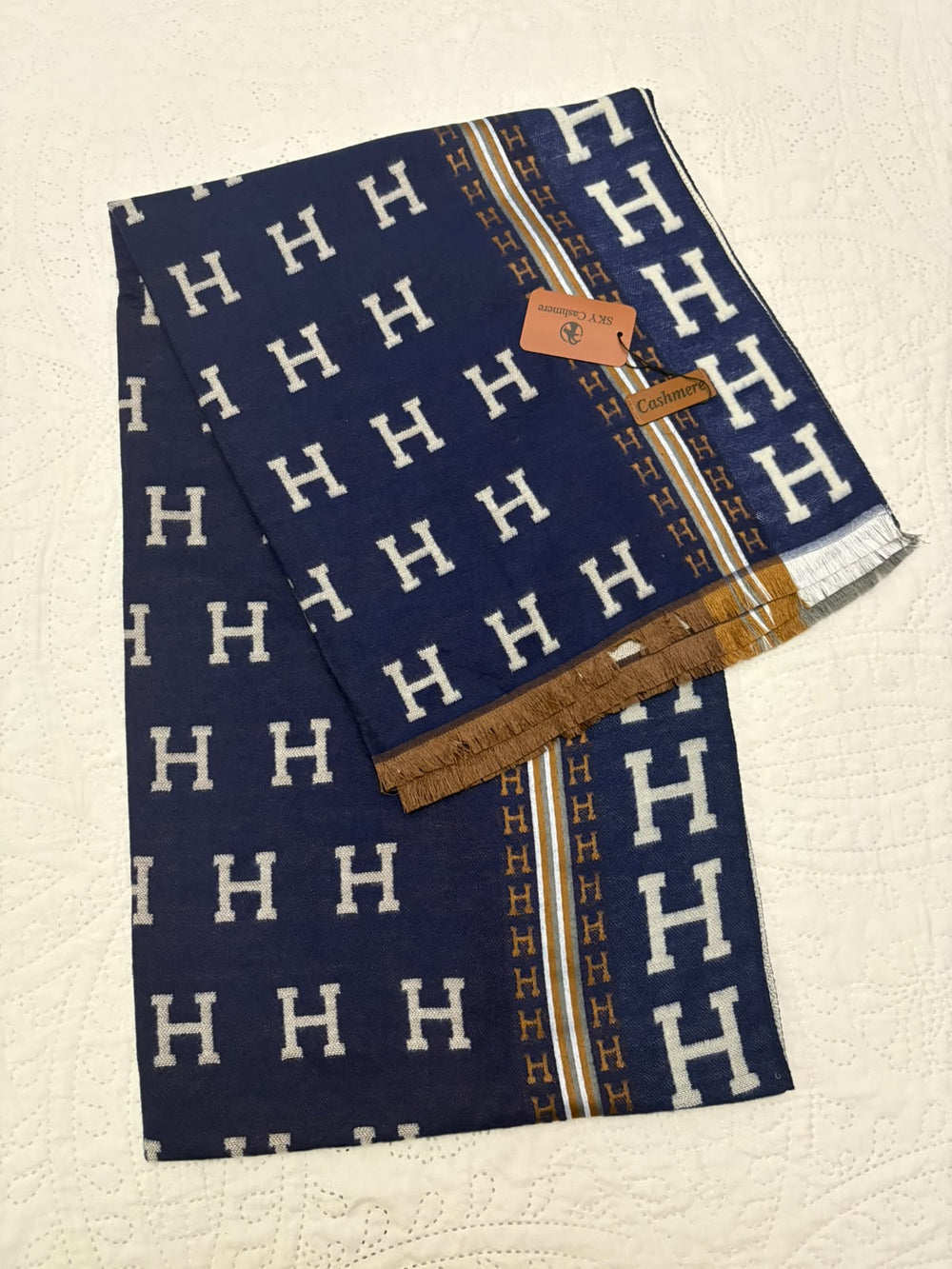 A folded navy blue scarf with a repeating white 'H' pattern, gold stripes, and a brown leather trim, displayed on a textured cream fabric background.