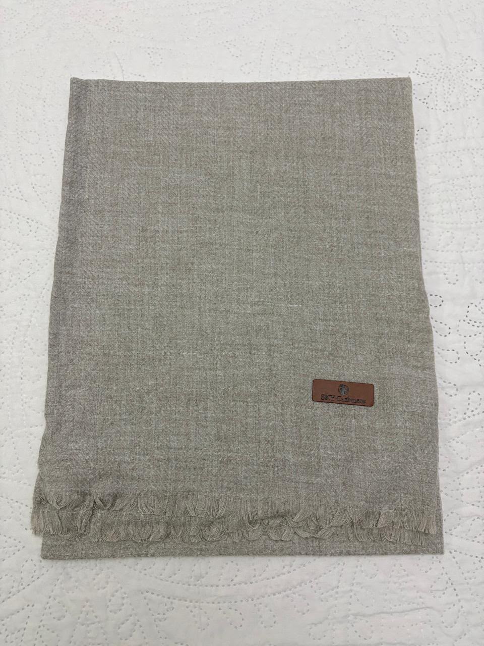 A folded light gray woven scarf with fringed edges and a small brown leather brand patch, laid flat on a white textured fabric background.