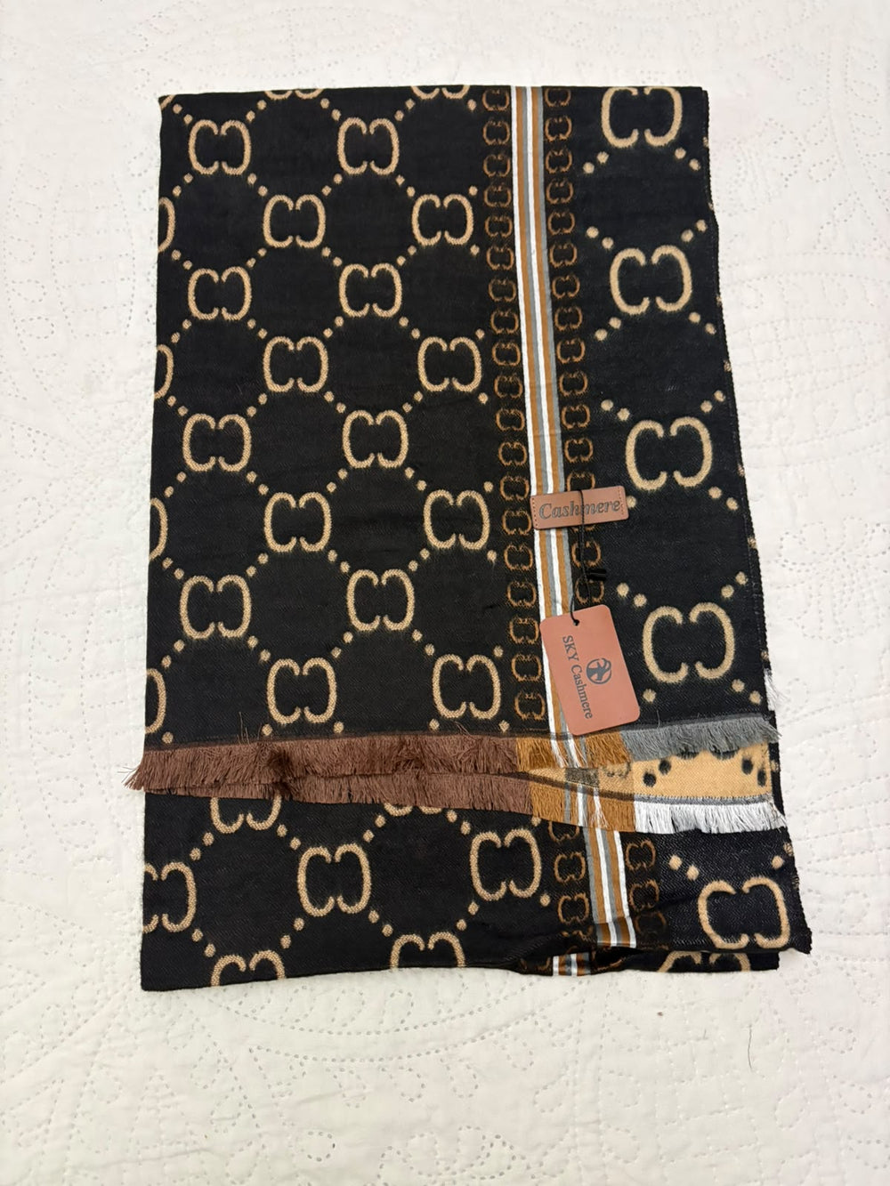 A folded black Gucci scarf featuring the signature beige GG monogram pattern, a vertical striped border with gold, brown, and white stripes, and fringed edges, displayed on a white textured fabric background.