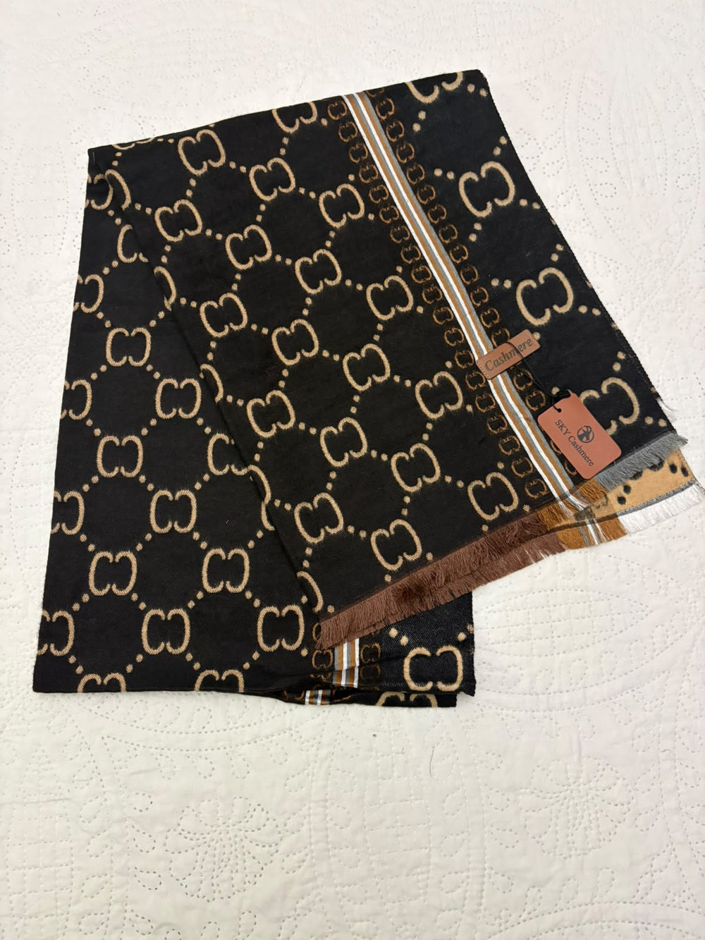 A folded black scarf featuring a beige interlocking GG monogram pattern, with a brown and beige striped border and a small leather Gucci brand tag.
