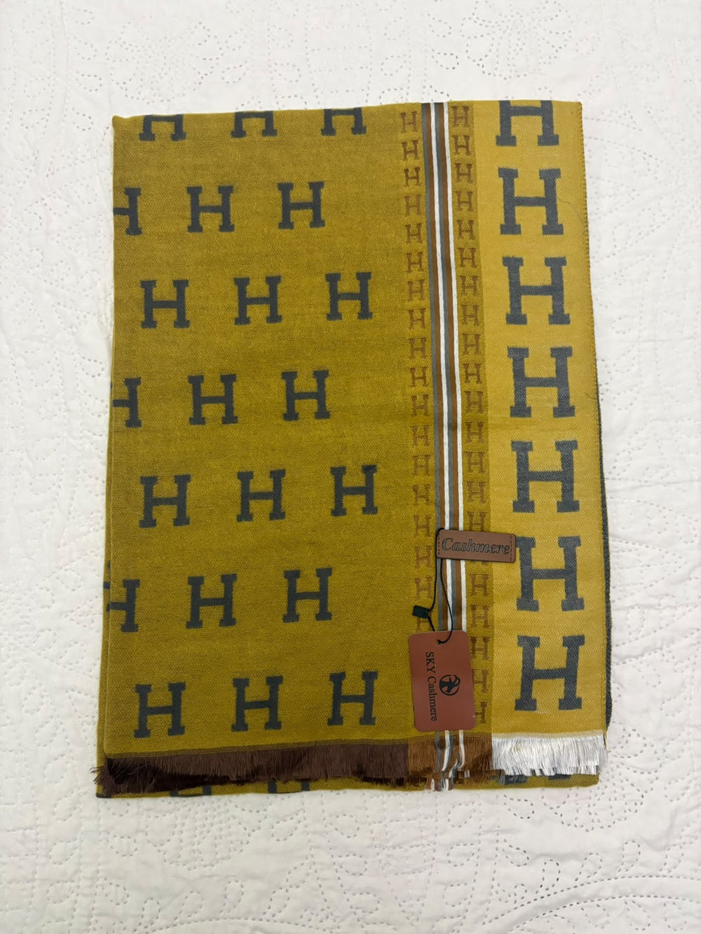 A folded yellow scarf with a repeating dark blue 'H' pattern, featuring a striped border and a brown leather brand tag that reads 'Hermès'.
