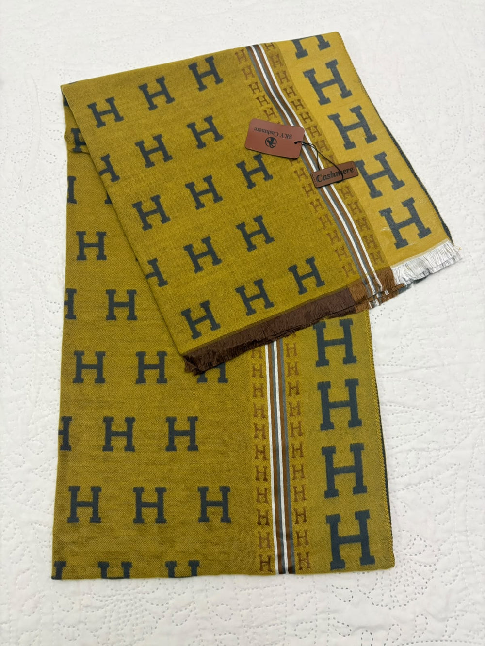A folded mustard yellow scarf with a repeating navy blue 'H' pattern, featuring a brown leather brand tag and fringed edges, displayed on a textured white fabric background.