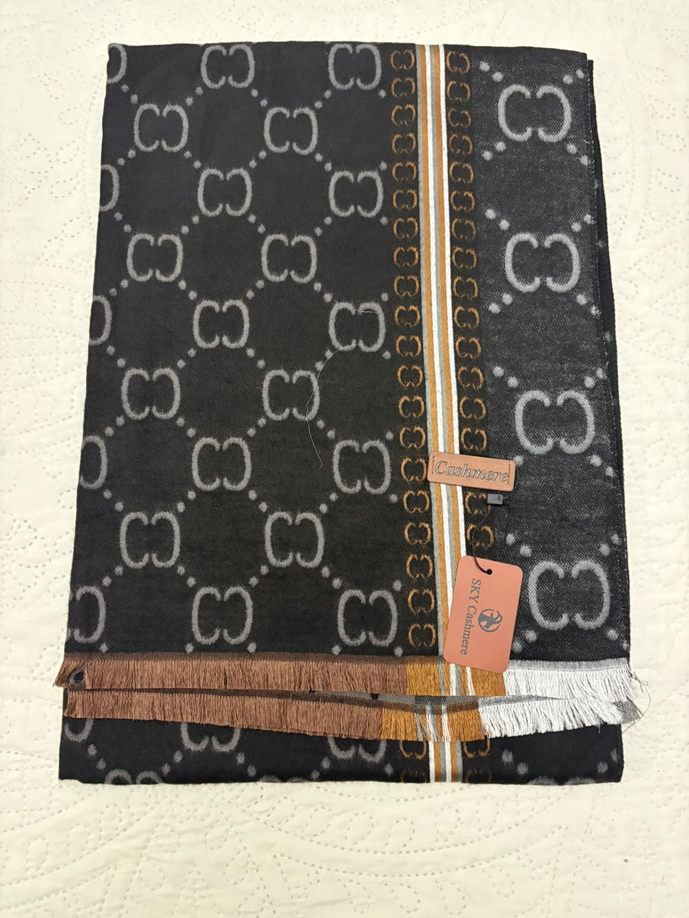 A folded Gucci scarf featuring a black base with a repeating white GG monogram pattern, a vertical gold and white stripe, and fringed edges, displayed on a textured off-white surface.