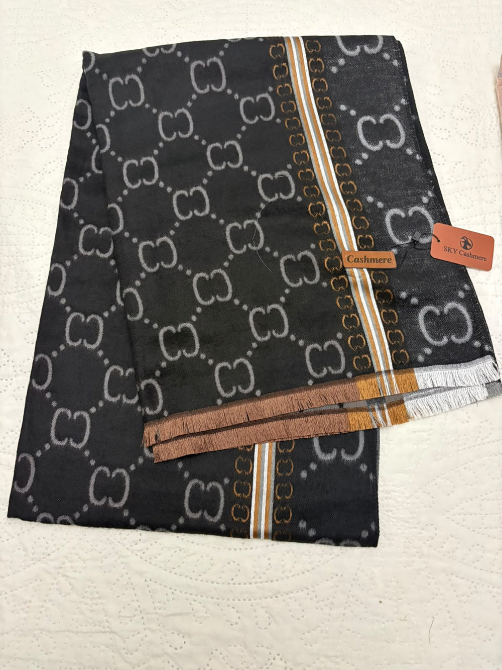 A folded black scarf featuring a repeating white monogram pattern, with decorative gold and white stripes, a brown fringe trim, and a small leather brand tag reading Cashmere. A matching orange tag is attached on the right side, and the scarf is laid flat on a white textured fabric background.
