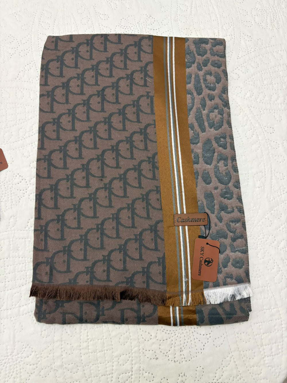 A folded designer scarf featuring a brown and gray monogram pattern on one side, a gray leopard print on the other, and a central gold stripe with thin white accents. A brown leather brand tag for 'Dior' is attached.