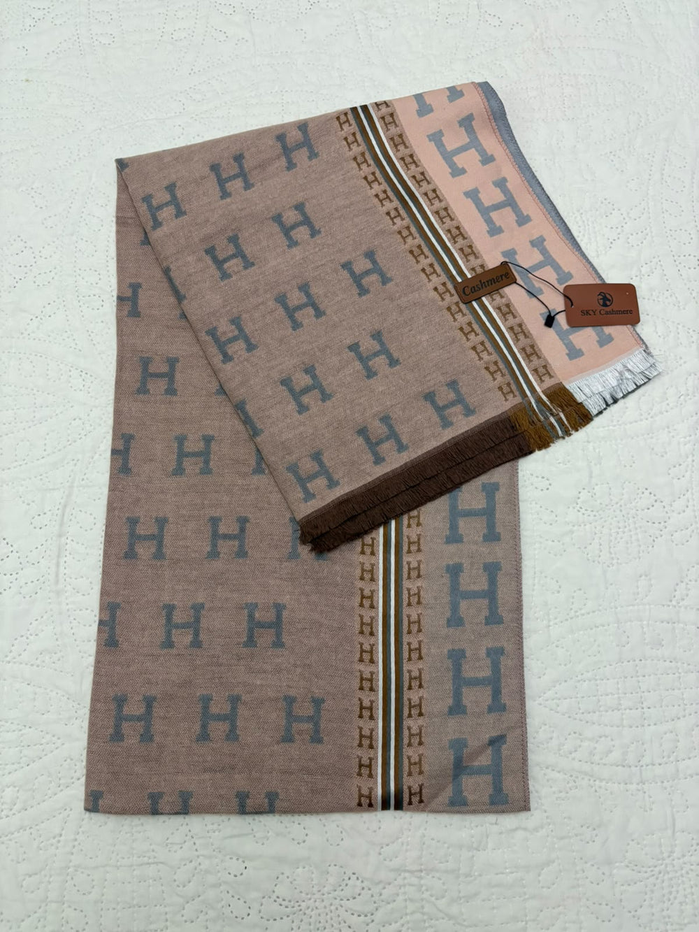 A folded Hermès scarf in taupe brown with a repeating blue 'H' pattern, a pink border with large blue 'H' motifs, and a brown leather brand tag attached, displayed on a textured white background.