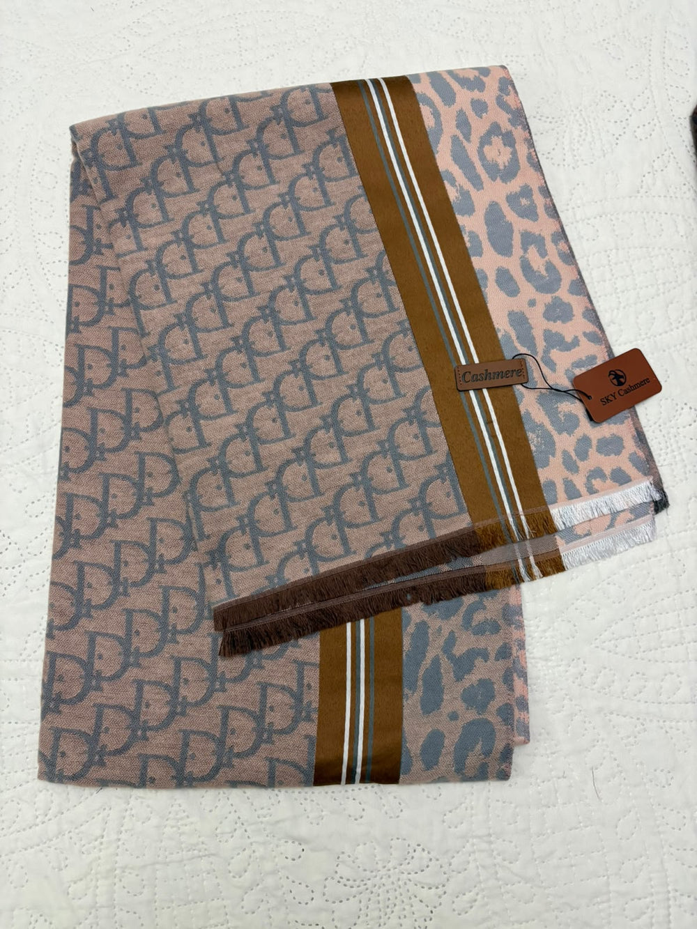 A folded designer scarf featuring a brown and gray repeating monogram pattern, a leopard print section, and a brown leather brand tag on a white textured background.