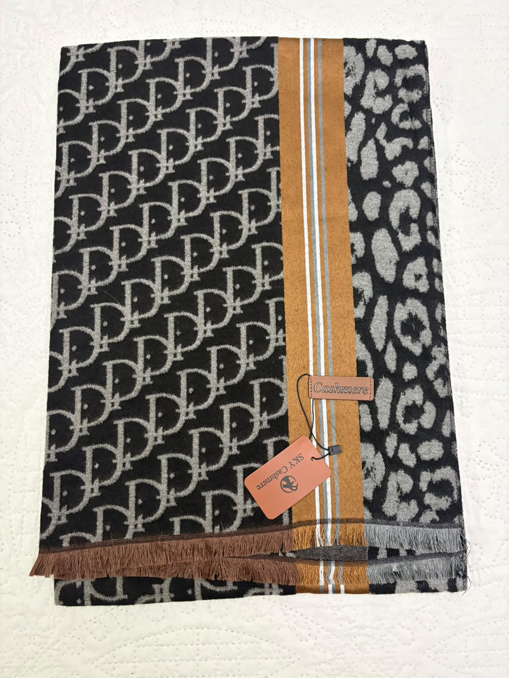 A folded designer scarf featuring a black and beige Dior Oblique monogram pattern on one side, a black and beige leopard print on the other, and a central vertical stripe panel in camel brown, white, and blue. A matching paper tag and a smaller orange hangtag are attached.