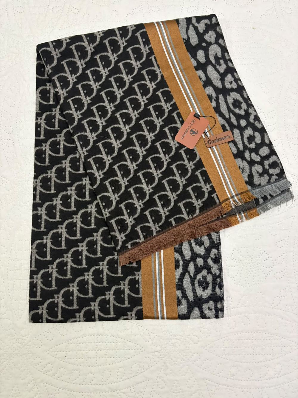 A folded black scarf featuring a repeating interlocking monogram pattern, a leopard print section, and a tan stripe with white double lines, displayed on a textured white surface with a brand tag attached.