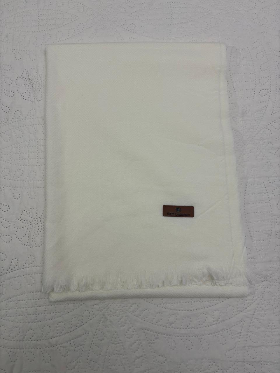 A folded white fringed scarf or throw blanket with a small brown leather brand tag, laid flat on a white textured fabric background.