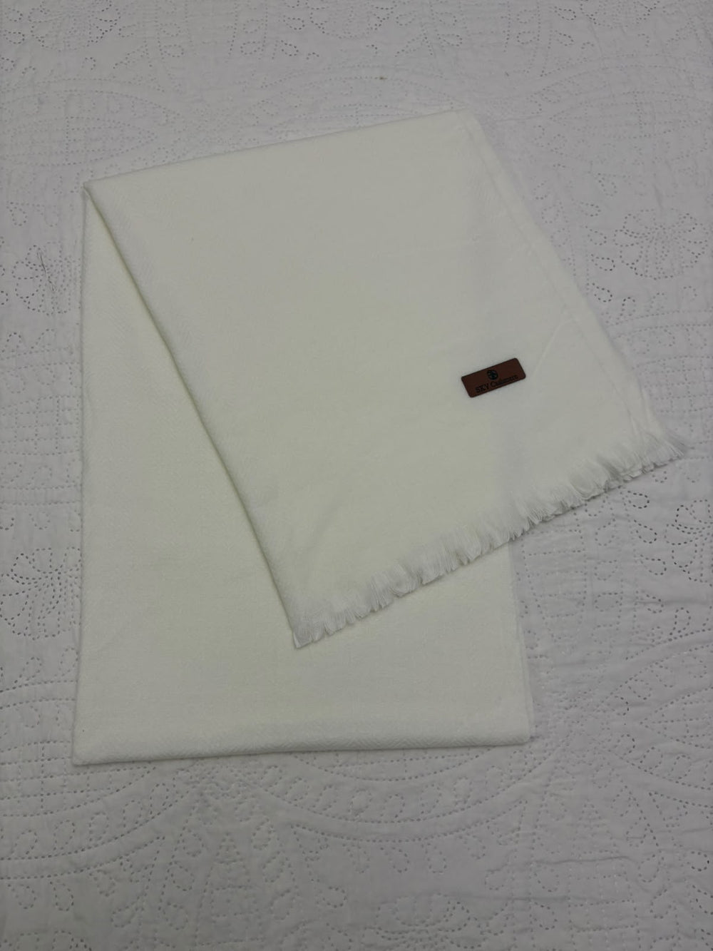 A folded off-white cotton throw blanket with fringed edges and a small brown brand tag, displayed on a textured white fabric background.