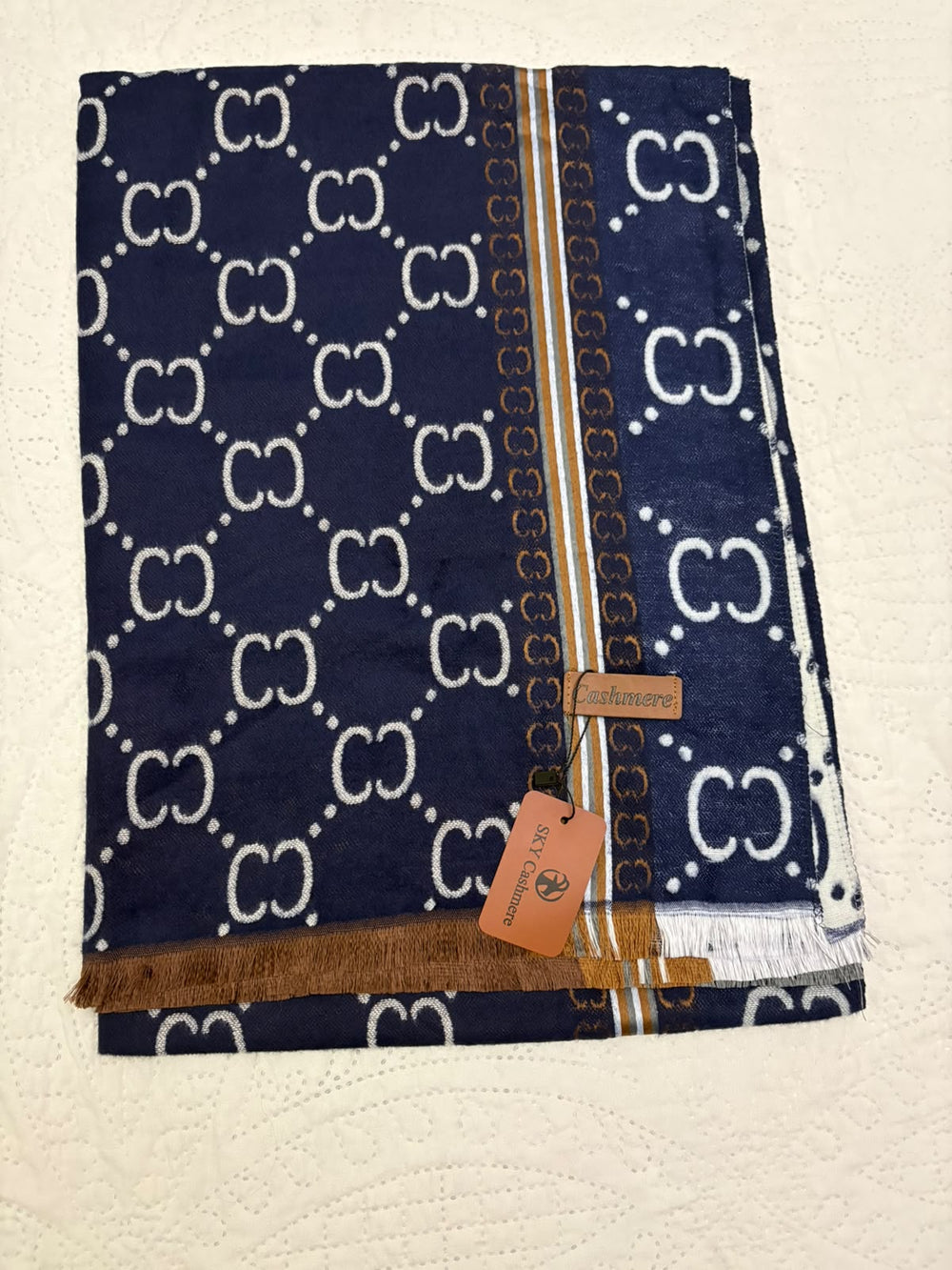 A folded navy blue Gucci scarf with a white GG monogram pattern, a gold and white border stripe, and a brown leather brand tag.