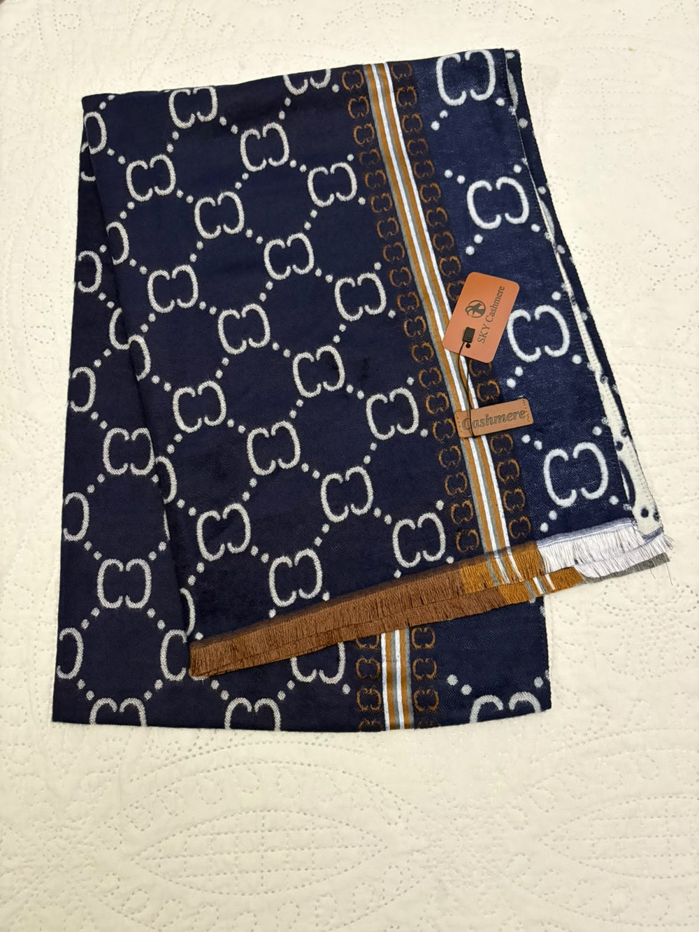 A folded navy blue scarf with a white interlocking CC monogram pattern, gold and white border stripes, and a small brown leather brand tag, displayed on a textured off-white surface.