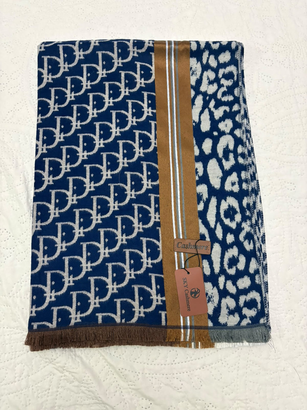 A folded navy blue scarf with a white repeating monogram pattern on one side, a white leopard print on the other, and a central tan stripe with thin white lines and fringe. A small paper tag is attached near the center.