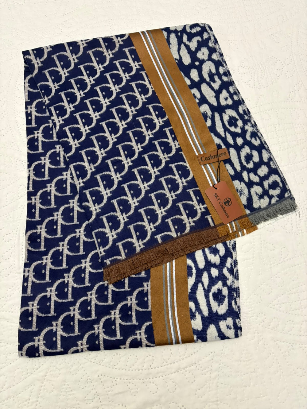 A folded navy blue scarf with a repeating white monogram pattern, a white leopard print section, and brown leather trim, displayed on a textured off-white surface.
