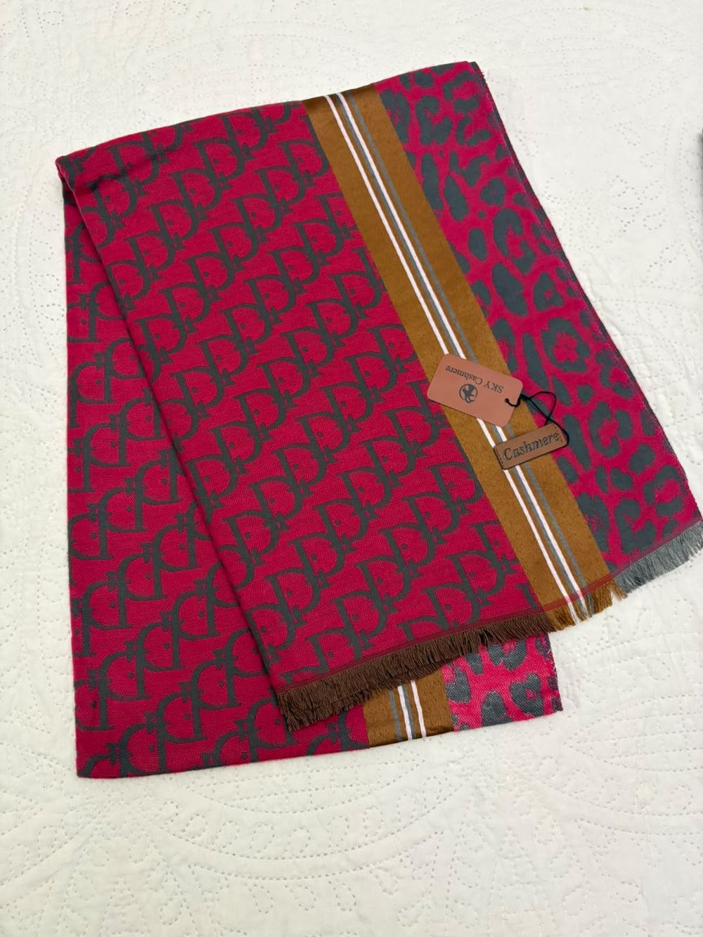 A folded pink scarf with a dark gray repeating monogram pattern, featuring a vertical gold stripe with white and silver lines and fringed edges. A small brand tag is attached, and the scarf is laid flat on a textured white surface.