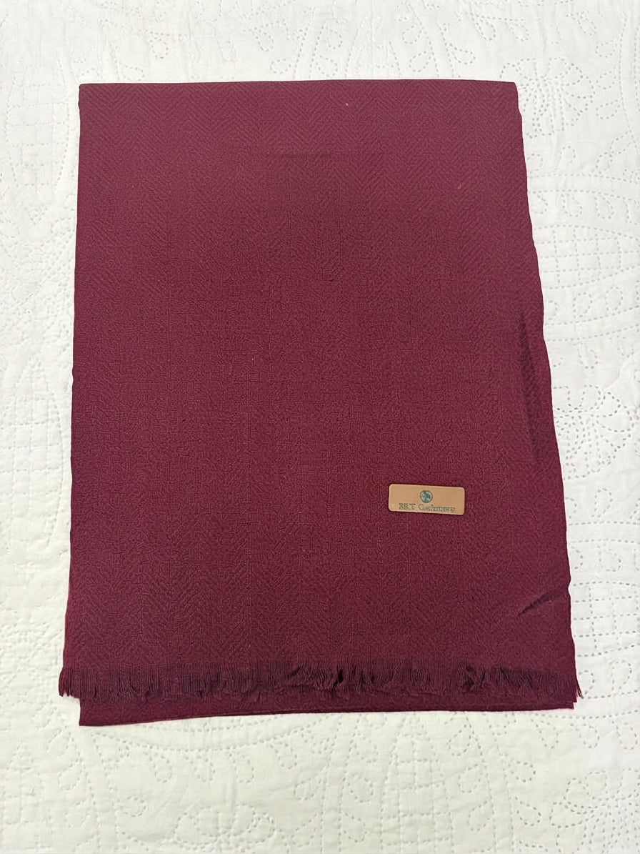 A folded deep burgundy scarf with a subtle woven texture and fringed edges, laid flat on a white textured fabric background. A small tan leather brand tag is attached near the lower right.