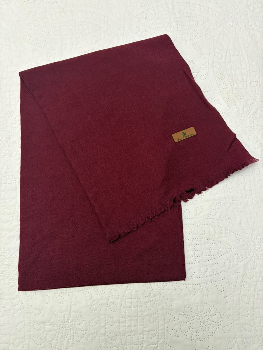 A folded deep burgundy scarf with a small tan leather brand tag, displayed on a textured white fabric background.
