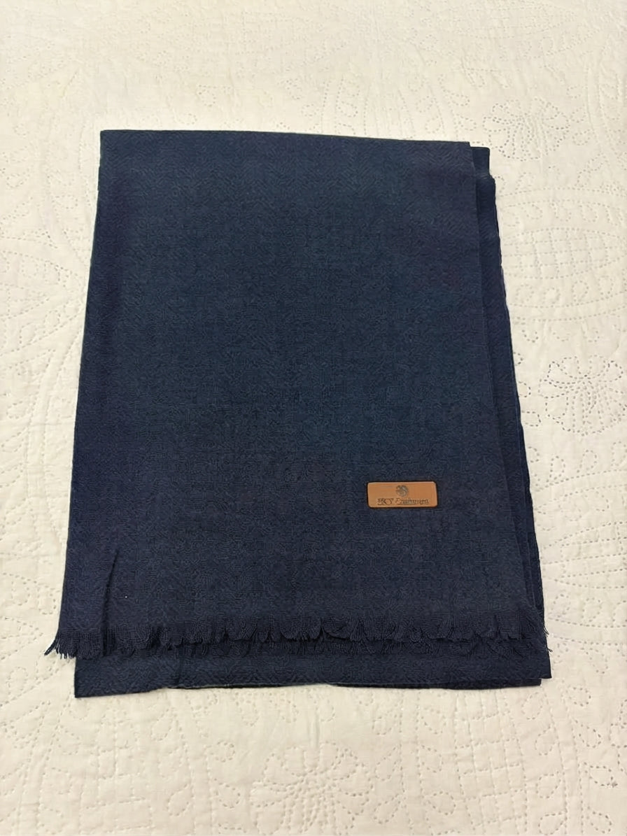 A folded navy blue scarf with fringed edges and a small tan leather brand label, placed on a textured off-white surface.