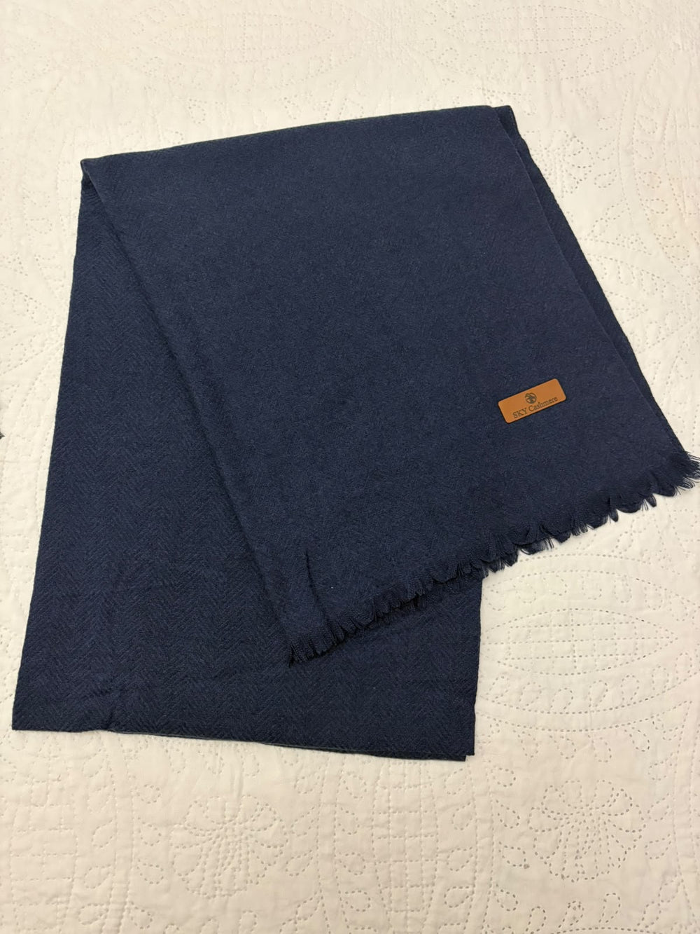 A folded navy blue scarf with fringed edges and a small tan leather brand tag, displayed on a textured off-white surface.