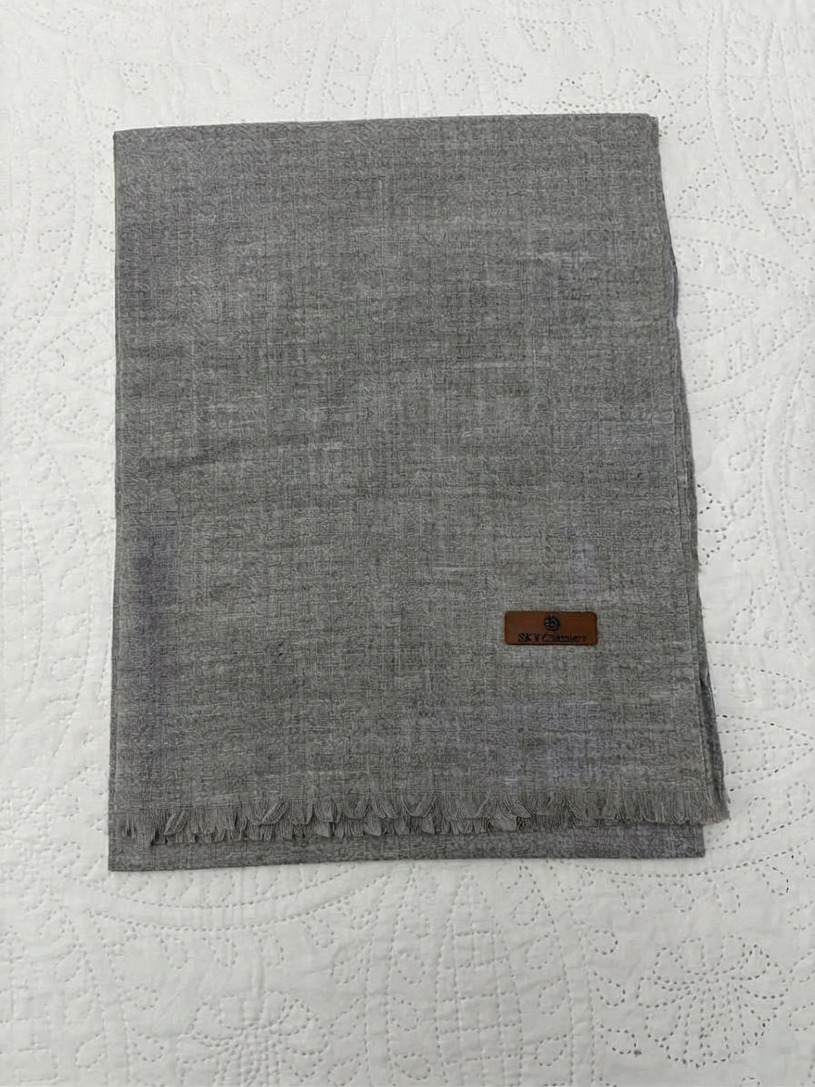 A folded gray woven scarf with fringed edges and a small brown leather brand patch, laid flat on a white textured fabric background.