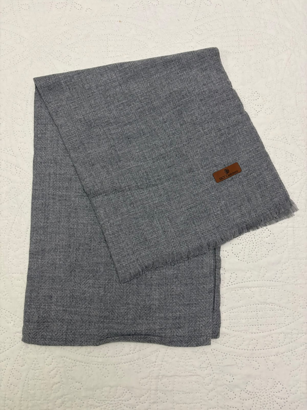 A folded gray woven scarf with a small brown leather brand tag, displayed on a textured white surface.
