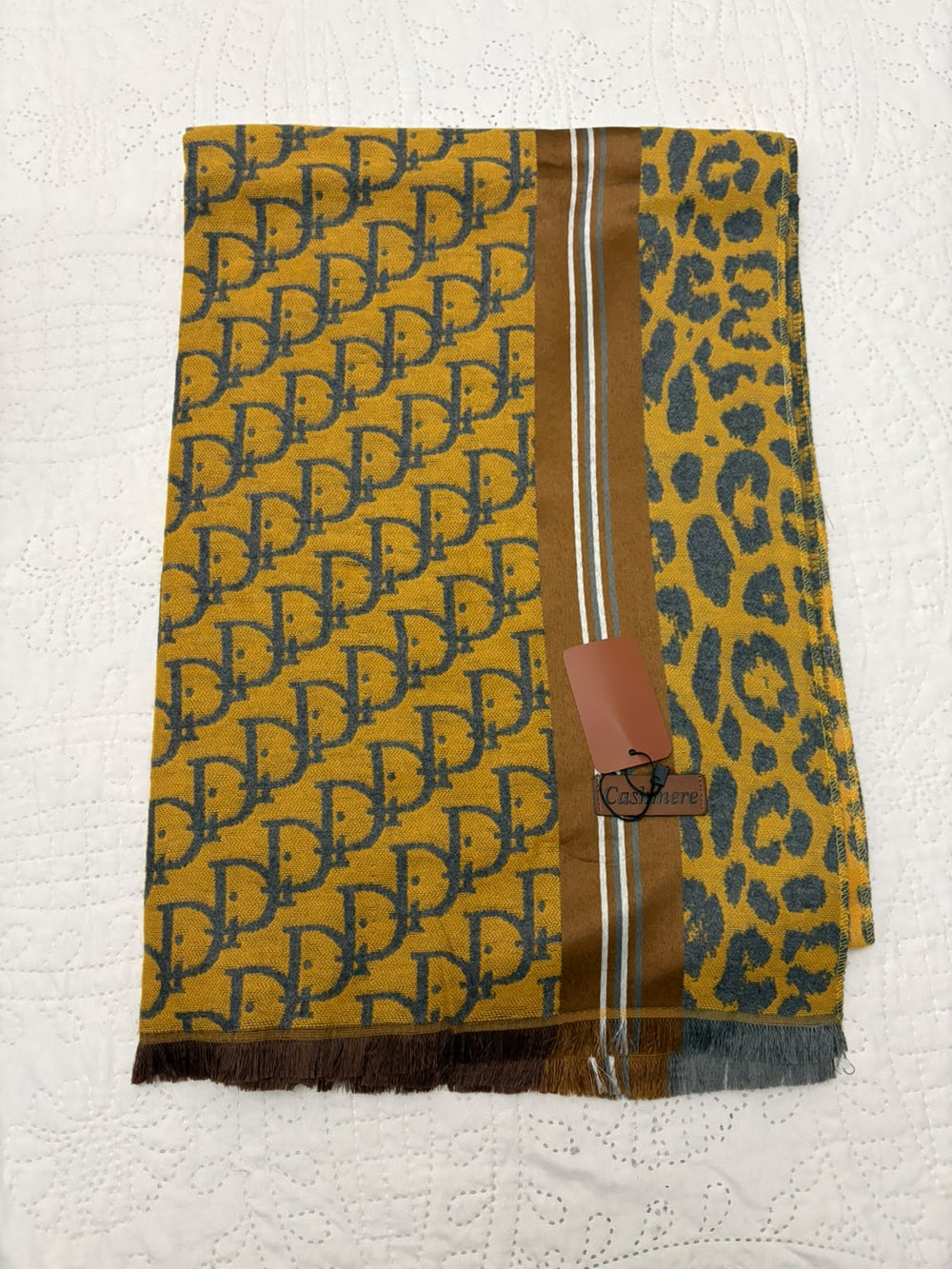 A folded designer scarf featuring a yellow and dark gray Dior Oblique pattern on one side, a dark gray leopard print on the other, and a brown center stripe with white accents. A brown leather tag is attached, and the scarf is laid flat on a white textured fabric background.