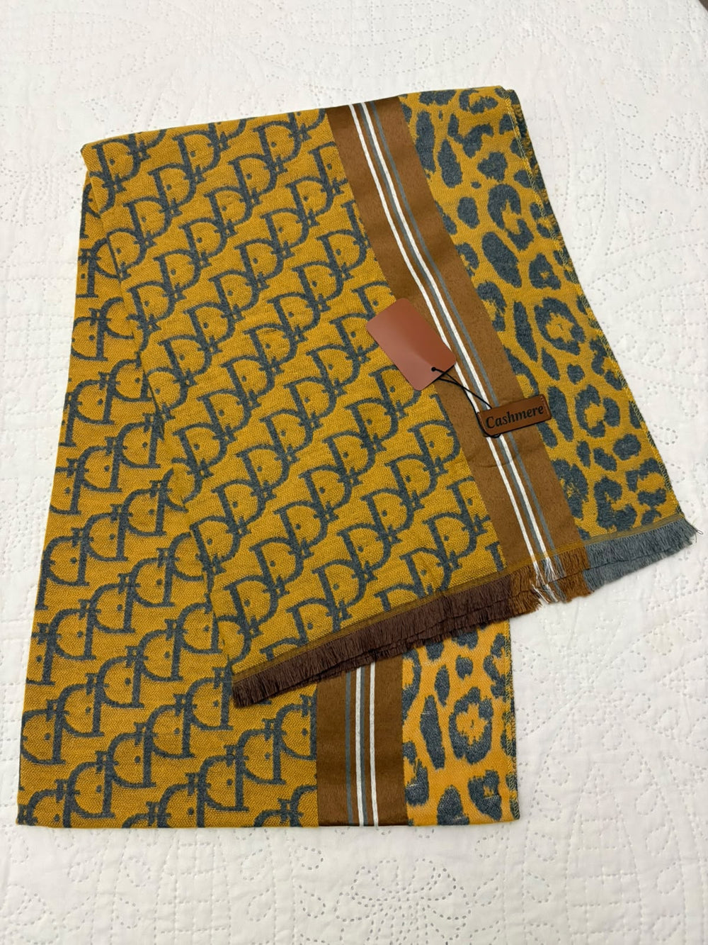A folded luxury scarf featuring a mustard yellow base with a dark blue repeating monogram pattern, a matching blue leopard print section, and brown stripes with white accents.