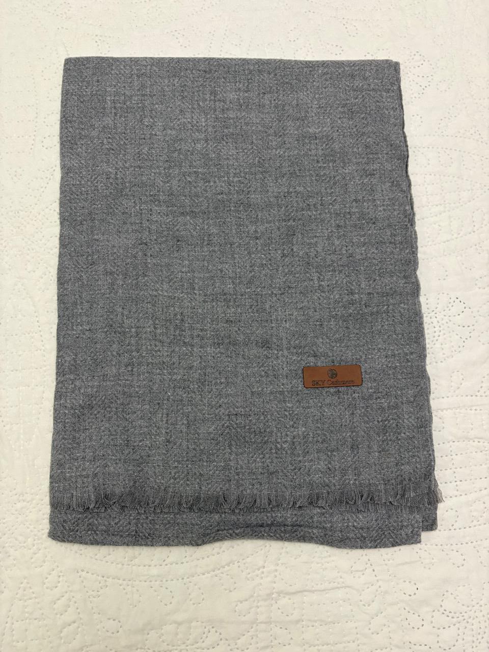 A folded gray woven scarf with fringed edges and a small brown leather brand tag, placed on a white textured fabric background.