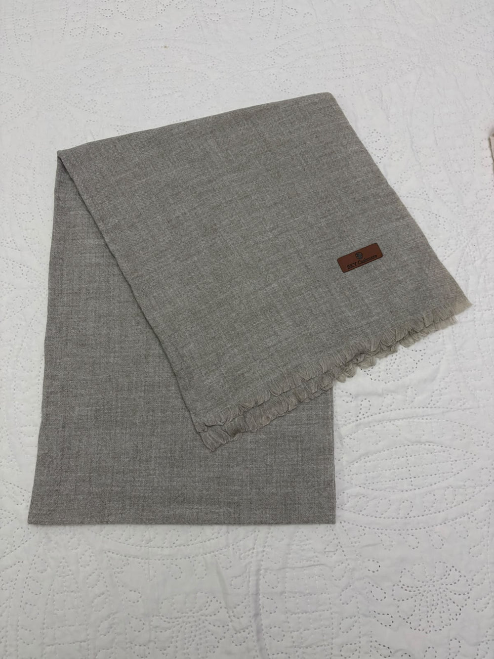 A folded gray woven scarf with fringed edges and a small brown leather brand tag, displayed on a white textured fabric background.