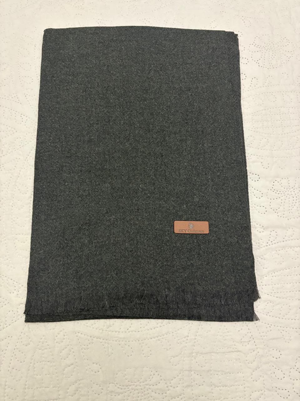 A folded dark gray wool scarf with a small brown leather brand patch, placed on a textured off-white surface.