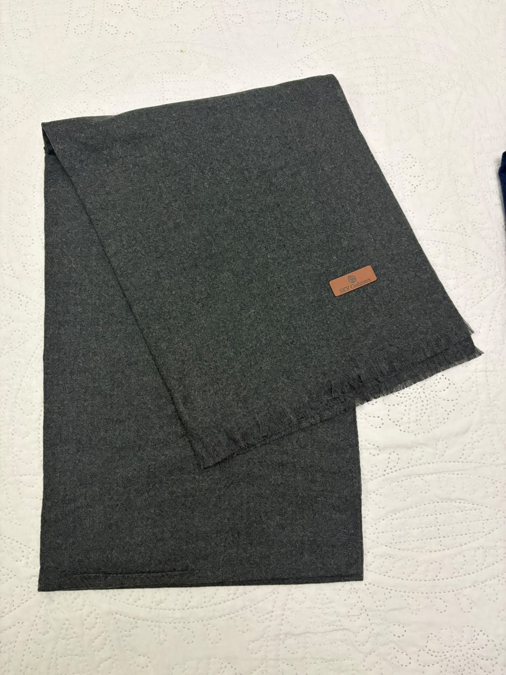 A dark charcoal gray folded scarf with a small brown leather brand tag, displayed on a textured white fabric background.