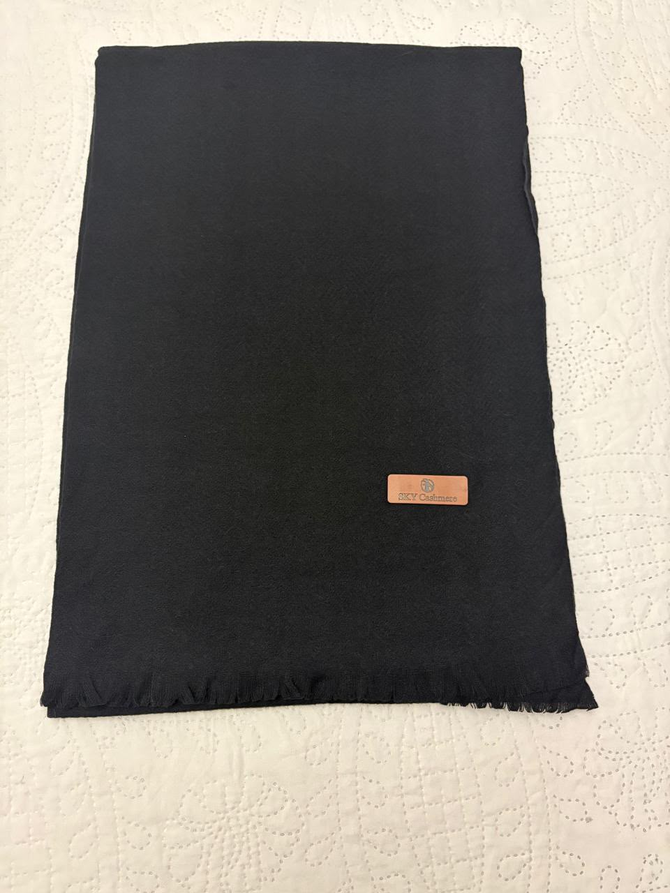 A folded black scarf with fringed edges and a small tan leather brand tag, laid flat on a textured off-white surface.