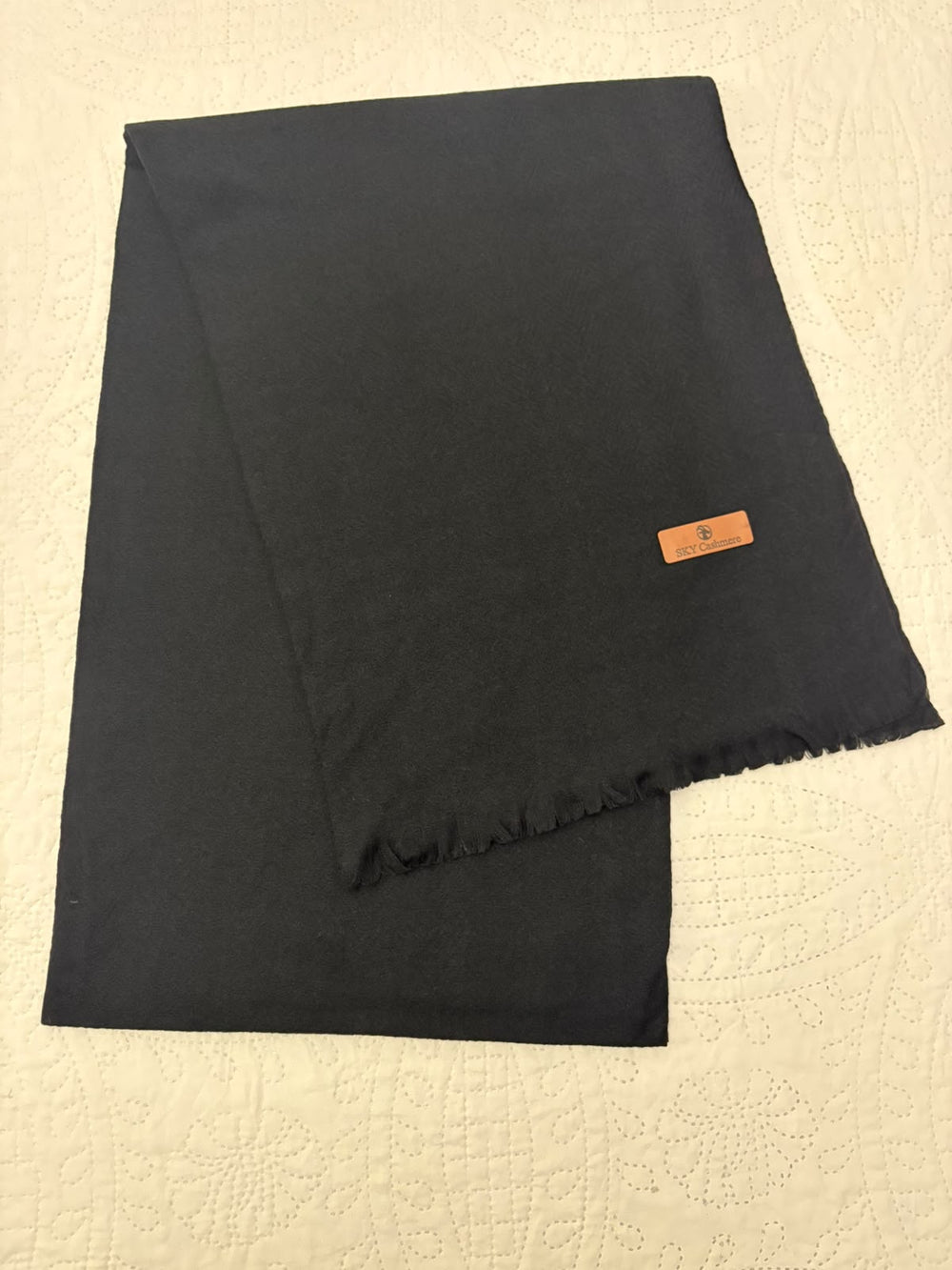 A folded black scarf with fringed edges and a small tan leather brand tag, laid flat on a textured cream-colored surface.